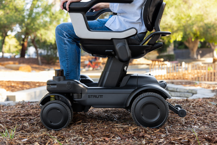 Strutt ev¹ smart power chair seen from the side on a garden path, showing the four wheels, joystick armrest and white seat shell