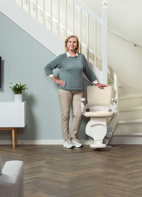 Otolift Modul-Air Smart curved stairlift parked at the foot of a white curved staircase, cream upholstered seat with armrest controls, a woman standing beside it.