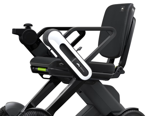 Close detail of the WHILL Model C Lite seat and controller: white arm-mounted joystick housing marked WHILL, padded backrest, and the carbon fibre cross-frame beneath the seat.