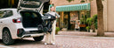 A person lifting the folded WHILL Model C Lite into the open boot of an estate car on a city street.